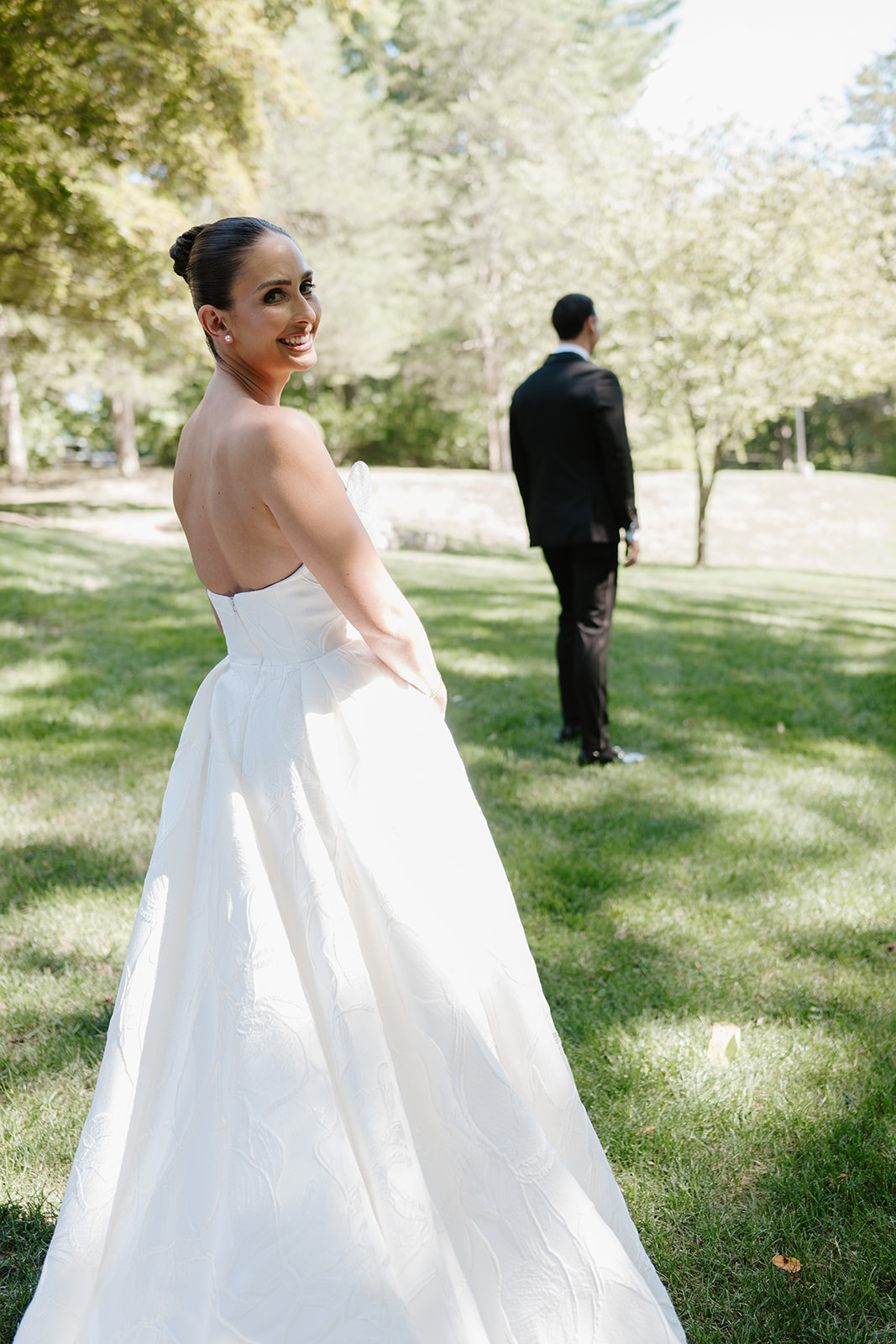 Smiling bride looking back at the camera as the groom stands with his back turned on a grassy field.